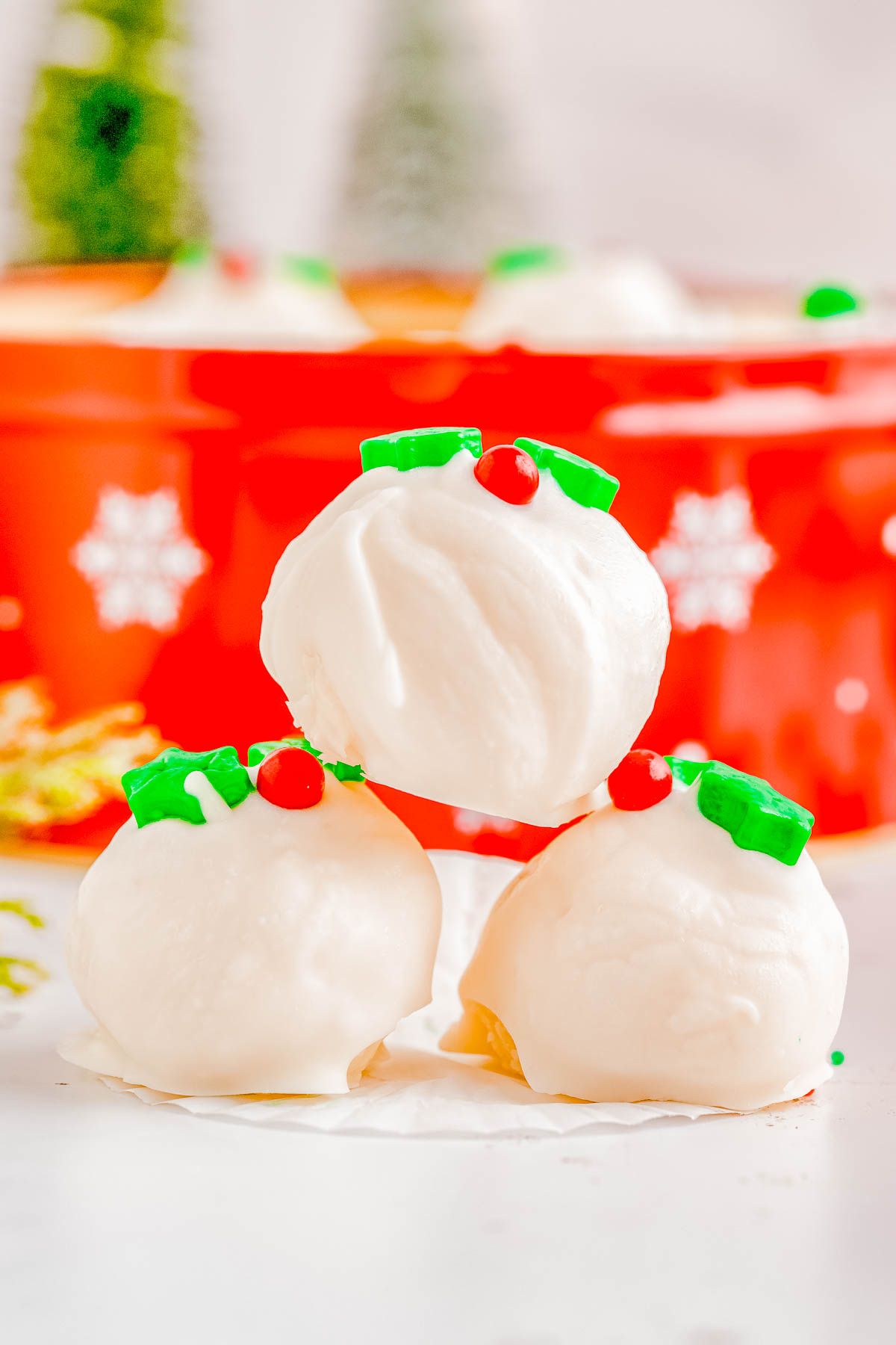 Three white chocolate-covered treats topped with green and red holiday decorations are arranged in front of a festive red container with snowflake patterns.