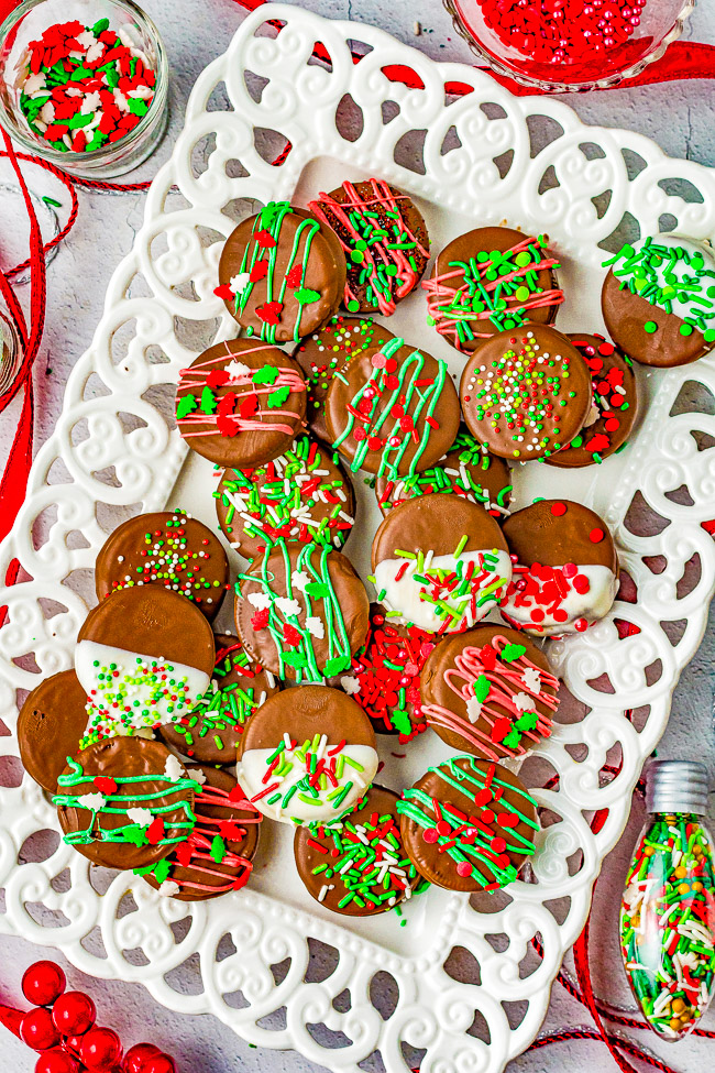 A white platter with chocolate-covered treats, decorated with red, green, and white icing and sprinkles. Surrounding are small bowls of sprinkles and ribbons.