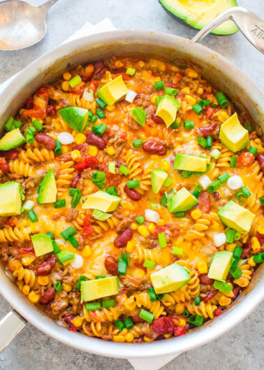 A skillet filled with a colorful pasta dish featuring rotini, beans, corn, diced tomatoes, melted cheese, and topped with avocado cubes and chopped green onions. An avocado half is seen beside the skillet.