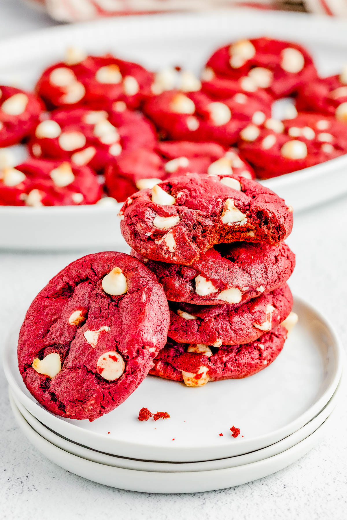 A stack of red velvet cookies with white chocolate chips on a plate, with more cookies in the background.