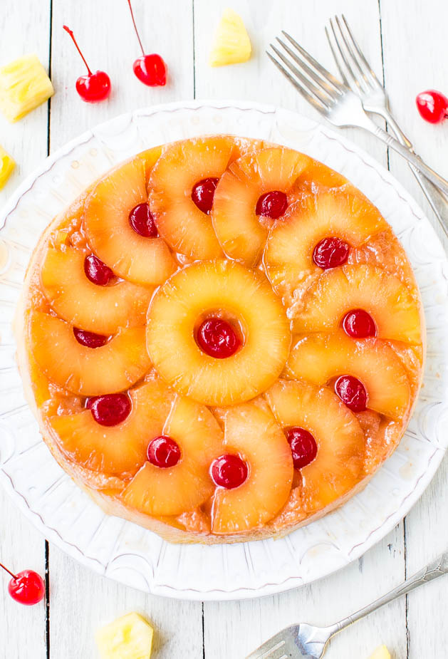A pineapple upside-down cake garnished with cherries, displayed on a white plate with forks on the side.