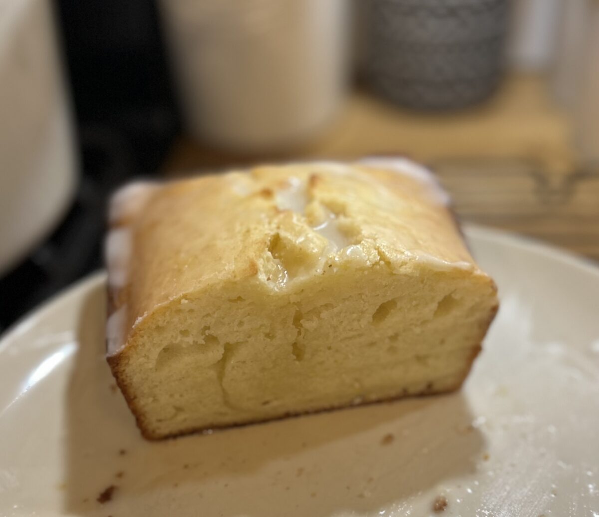 A loaf of lemon cake with a slice cut off, resting on a plate. The cake shows a moist, dense texture.