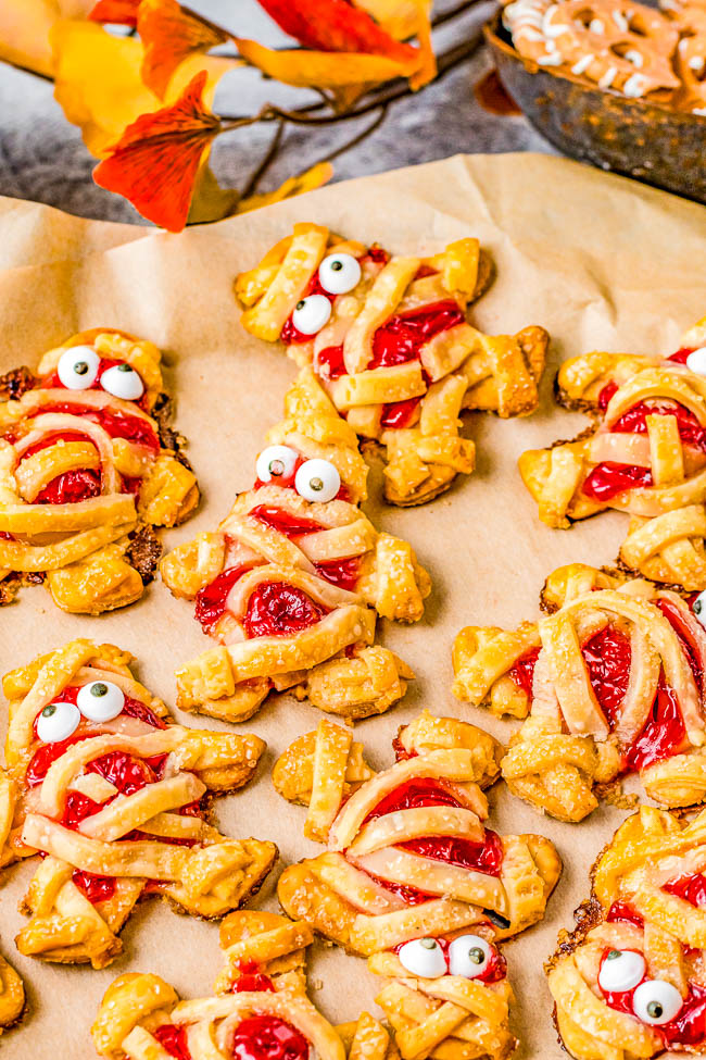 Cookies shaped like mummies with red filling and candy eyes are displayed on parchment paper. Orange leaves are in the background.