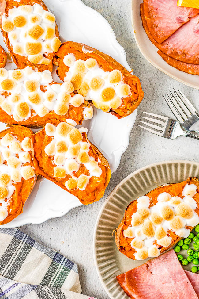 A plate with five sweet potato halves topped with toasted marshmallows, next to a plate containing slices of ham and peas. A striped napkin and two forks are also visible.