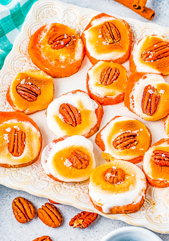 A decorative white plate holds a variety of baked sweet potato slices topped with melted marshmallows, pecan halves, and a sprinkle of coarse salt. Several loose pecans are scattered nearby.