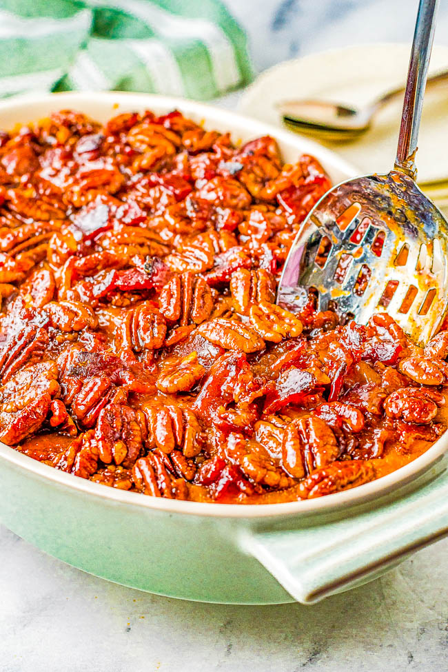 A close-up of a casserole dish filled with pecan-topped baked sweet potatoes. A serving spoon rests in the dish.