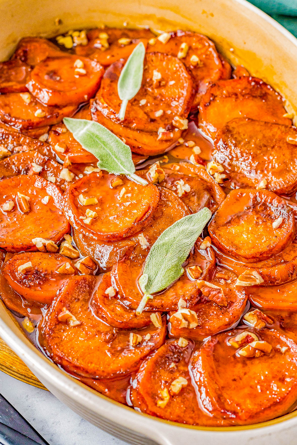 A close-up of a dish of candied sweet potatoes garnished with whole sage leaves and chopped nuts.