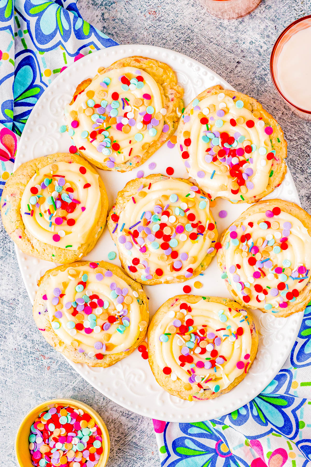 A white plate with seven frosted cookies covered in colorful sprinkles, placed on a table with a patterned tablecloth and a small bowl of sprinkles nearby.