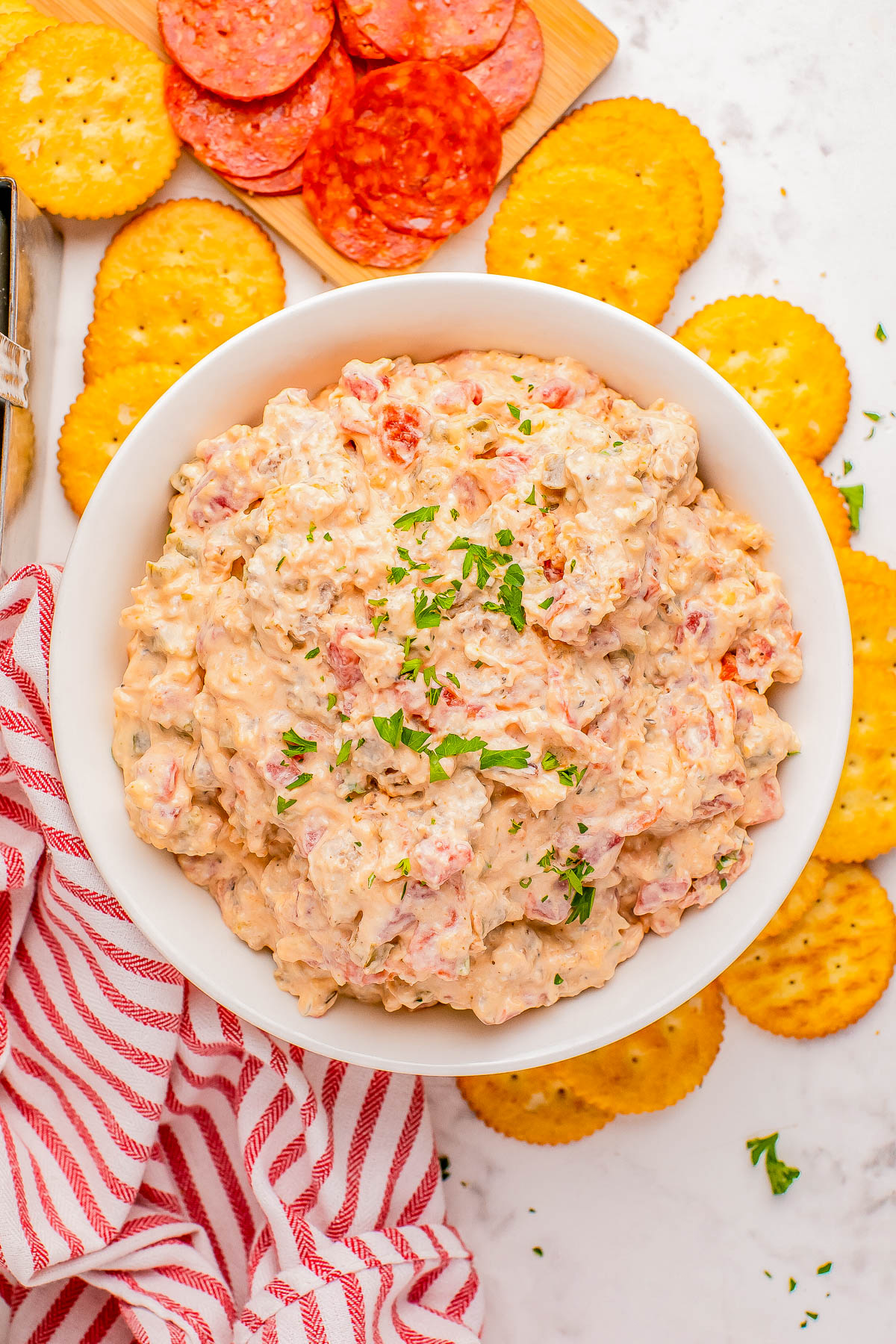 A bowl of creamy, textured cheese dip garnished with parsley, surrounded by round crackers and slices of salami on a marble surface with a red striped napkin.