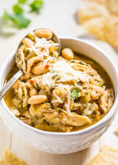 A bowl of white chicken chili topped with shredded cheese and garnished with peanuts, with a spoon inserted in the bowl.