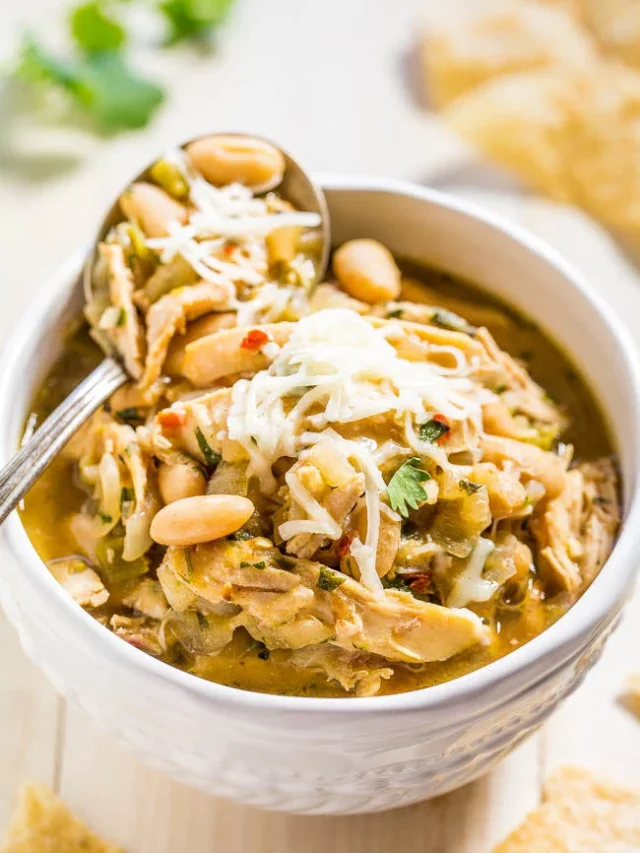 A bowl of white chicken chili topped with shredded cheese and garnished with peanuts, with a spoon inserted in the bowl.