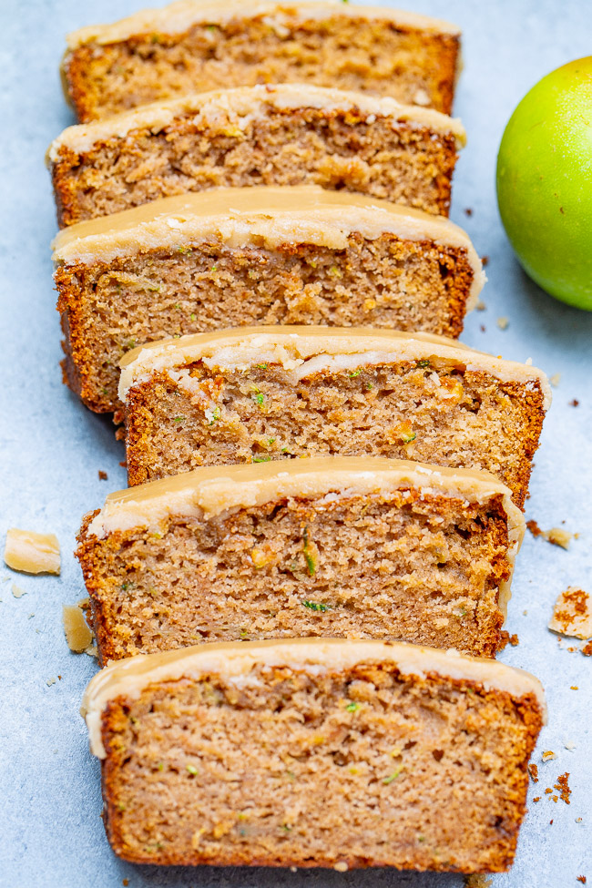 Slices of zucchini bread with frosting arranged in a line on a light gray surface beside a green apple.