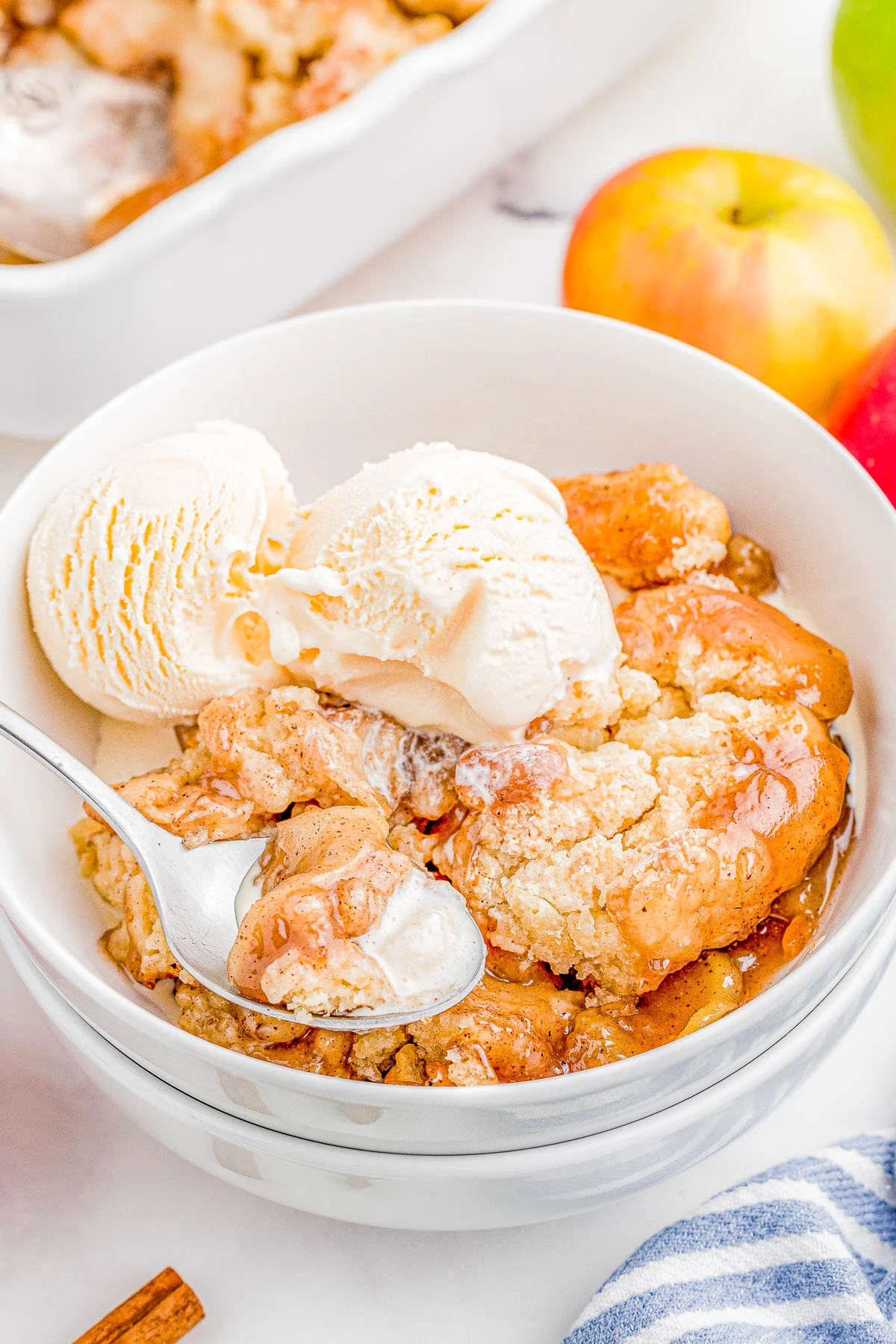 A bowl of apple crumble topped with vanilla ice cream. A spoon is holding a bite of the dessert. An apple and a baking dish with more crumble are in the background.