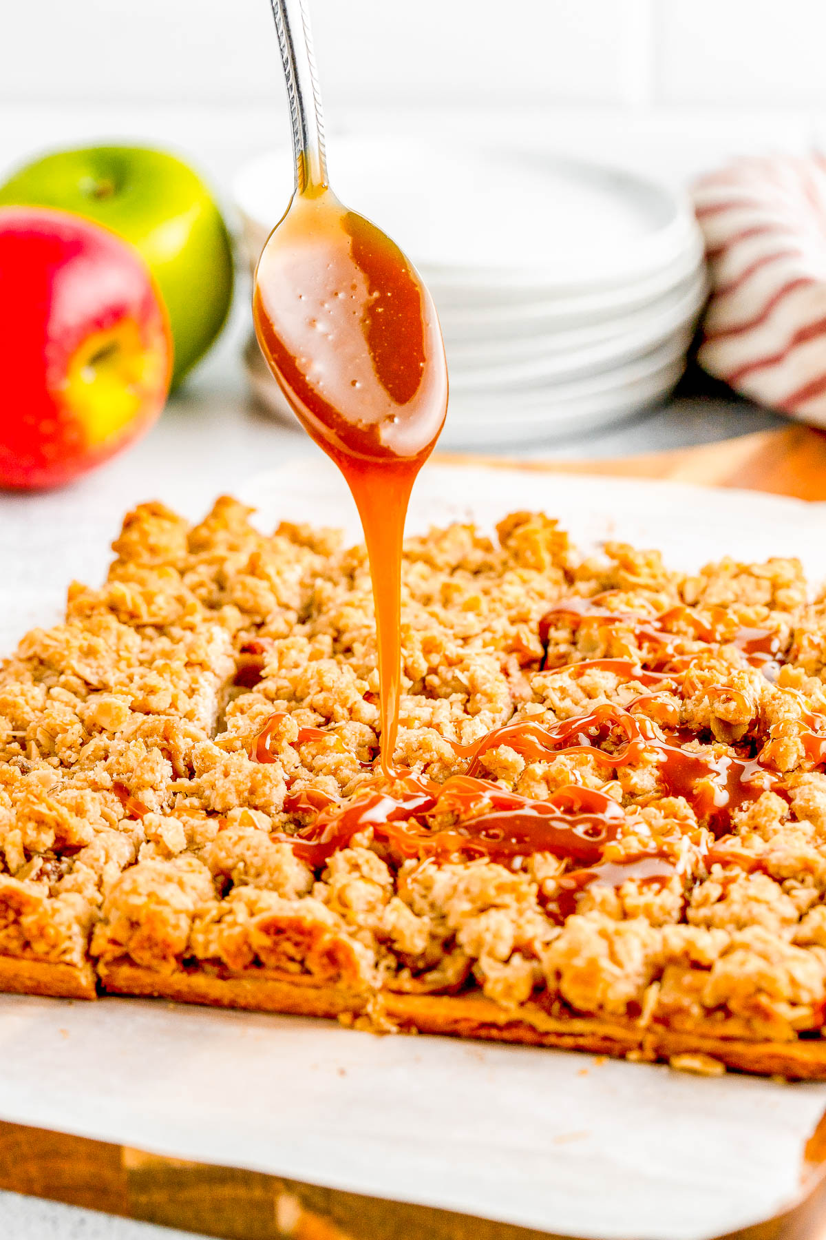 Caramel is being drizzled over a freshly baked crumb cake, with a spoon hovering above. In the background, there are stacked plates, a striped cloth, and two apples.