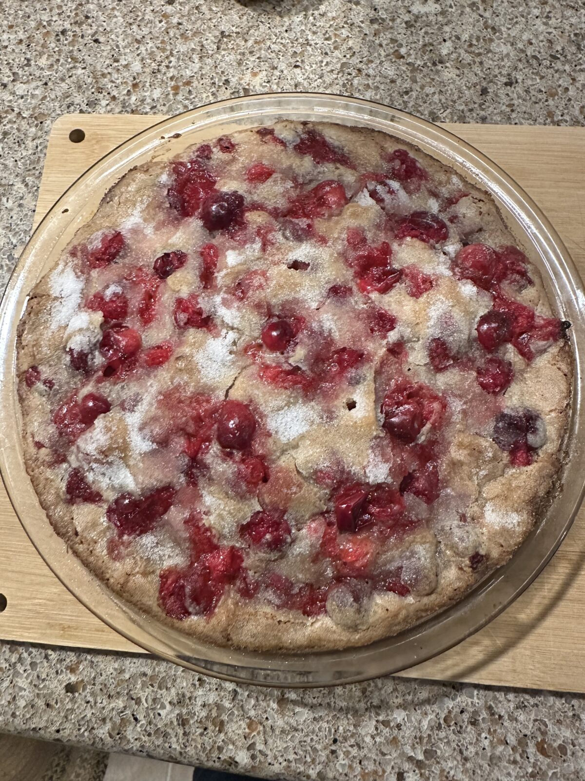 A round, freshly baked pie with a golden crust topped with scattered red berries and a dusting of sugar, placed on a wooden board.