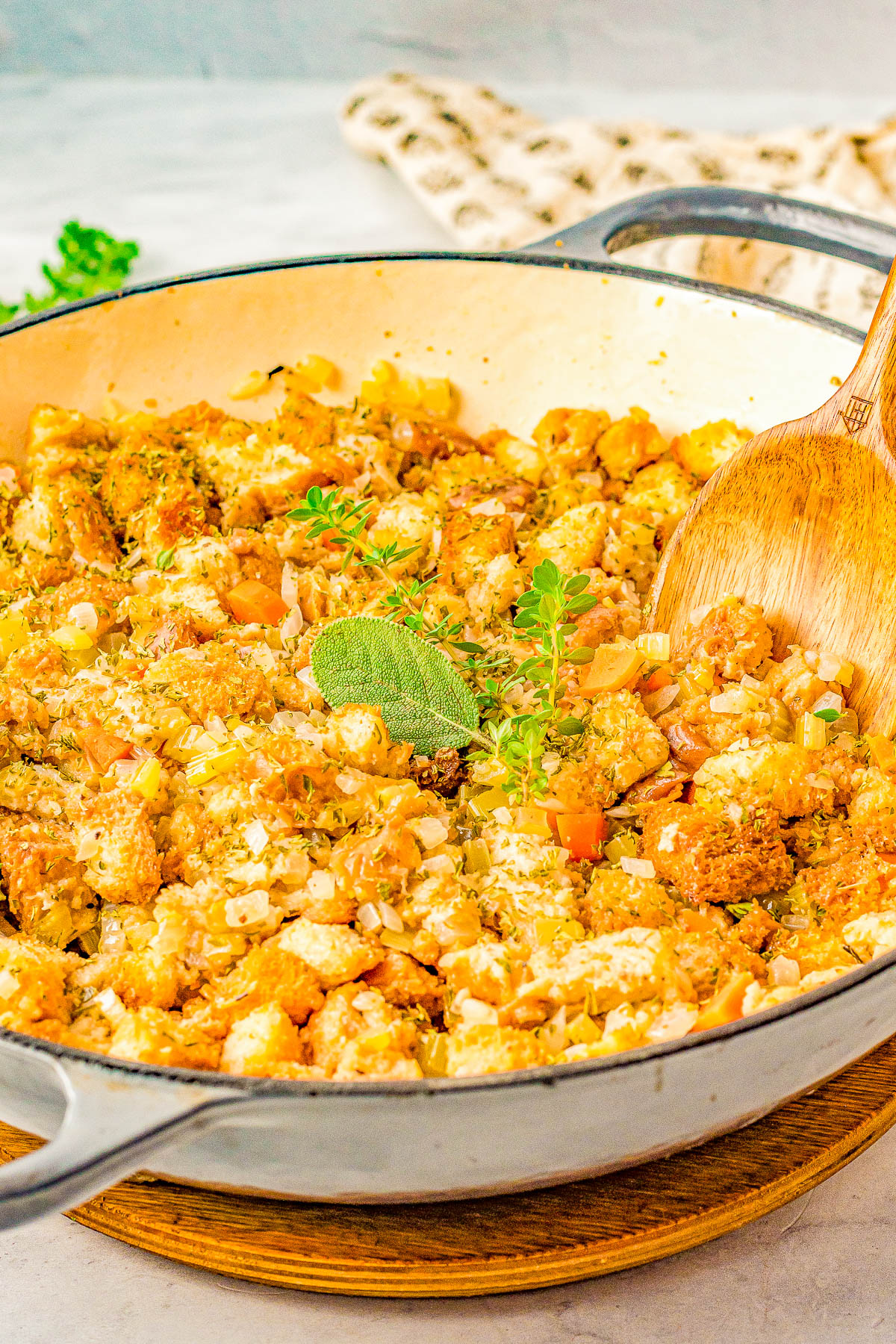 A skillet containing a cooked mixture of breadcrumbs, onions, celery, and herbs, with a wooden spoon and a sprig of fresh herbs on top.