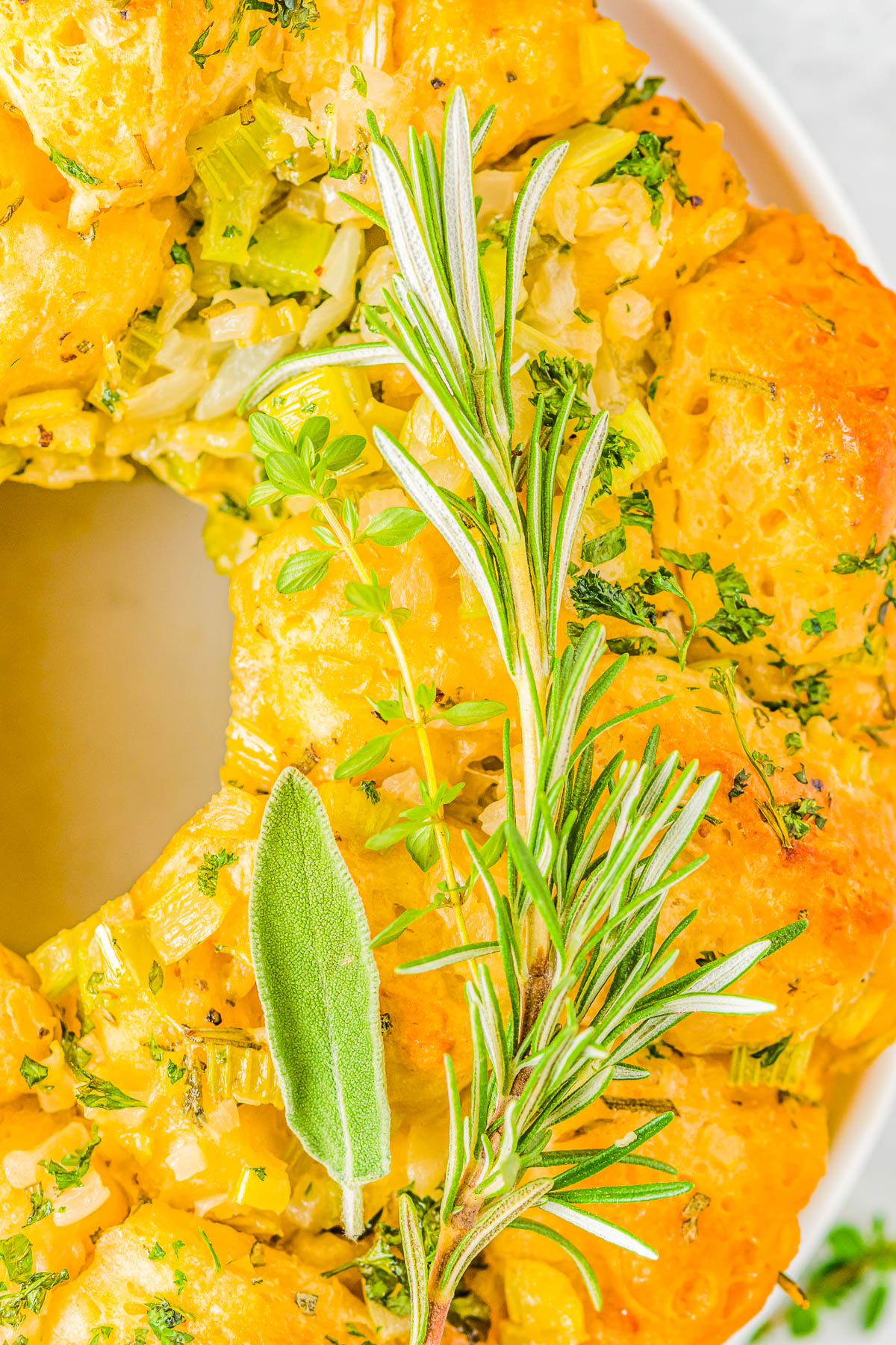 Close-up of a golden-brown savory pull-apart bread ring garnished with fresh rosemary, parsley, and sage herbs, displayed on a white plate.