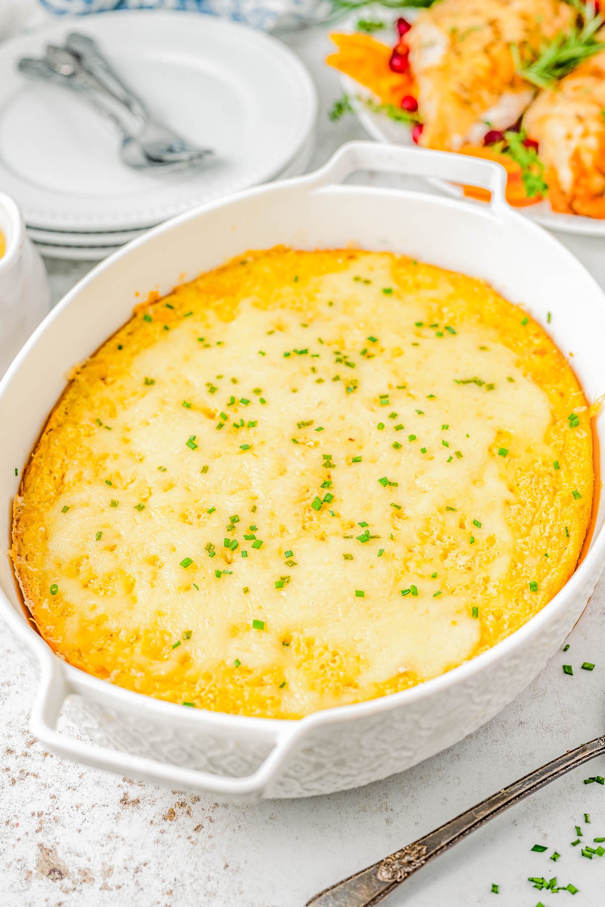 A baked yellow dish topped with melted cheese and garnished with chopped green herbs in a white oval dish on a table. Plates and utensils are in the background.