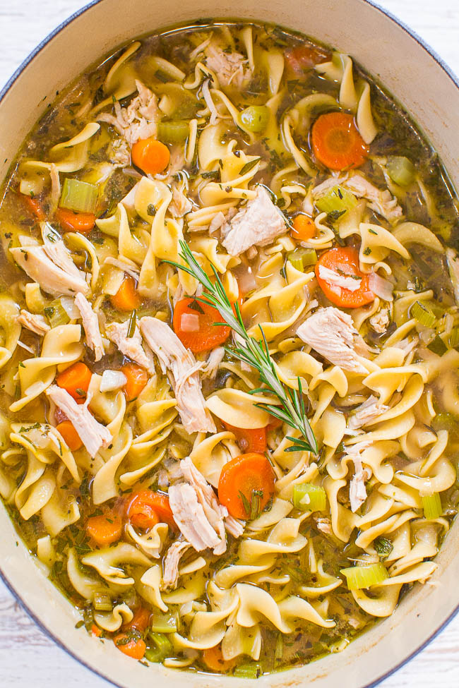 A pot of chicken noodle soup containing chunks of chicken, egg noodles, sliced carrots, celery, and garnished with a rosemary sprig.