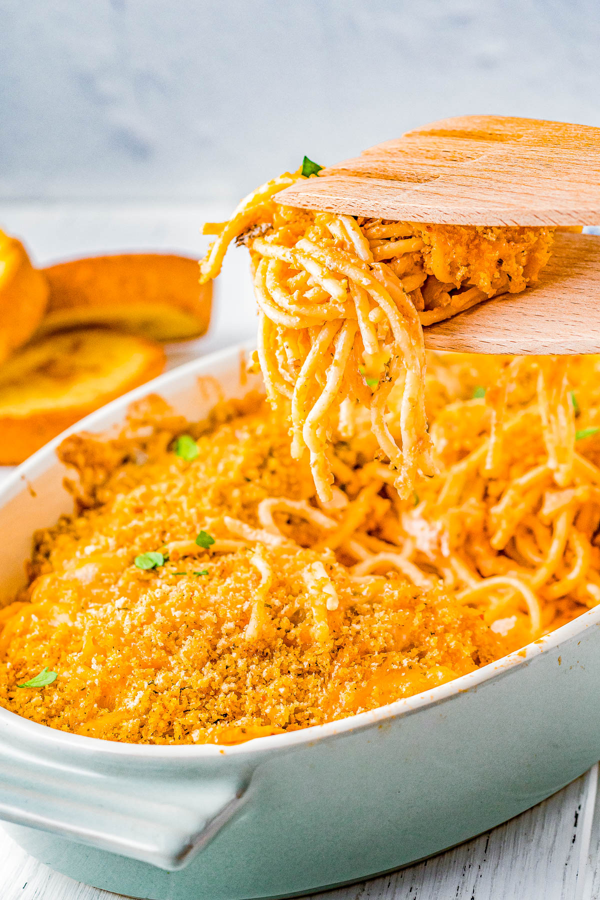 A wooden spatula is lifting a portion of baked spaghetti coated with a golden breadcrumb topping from a casserole dish. A sliced squash is visible in the background.