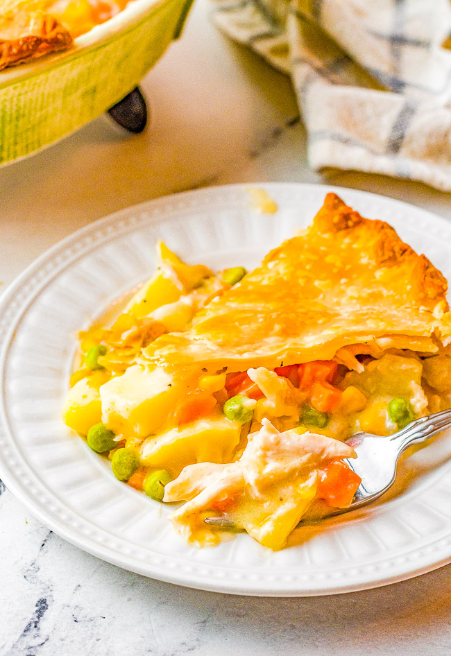 A slice of chicken pot pie on a white plate, with a visible filling of vegetables including peas, carrots, and potatoes, and a golden-brown crust. A fork rests beside the slice.