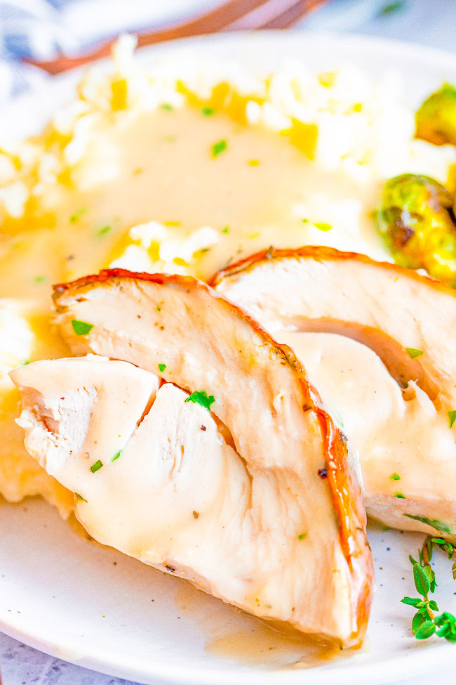 A plate of sliced roasted chicken breast with gravy, served alongside mashed potatoes and roasted Brussels sprouts.