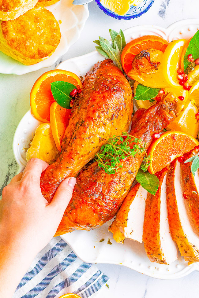 A hand reaching towards roasted turkey legs and sliced breast meat garnished with orange slices and herbs on a white plate. Nearby, biscuits and a striped cloth are partially visible.