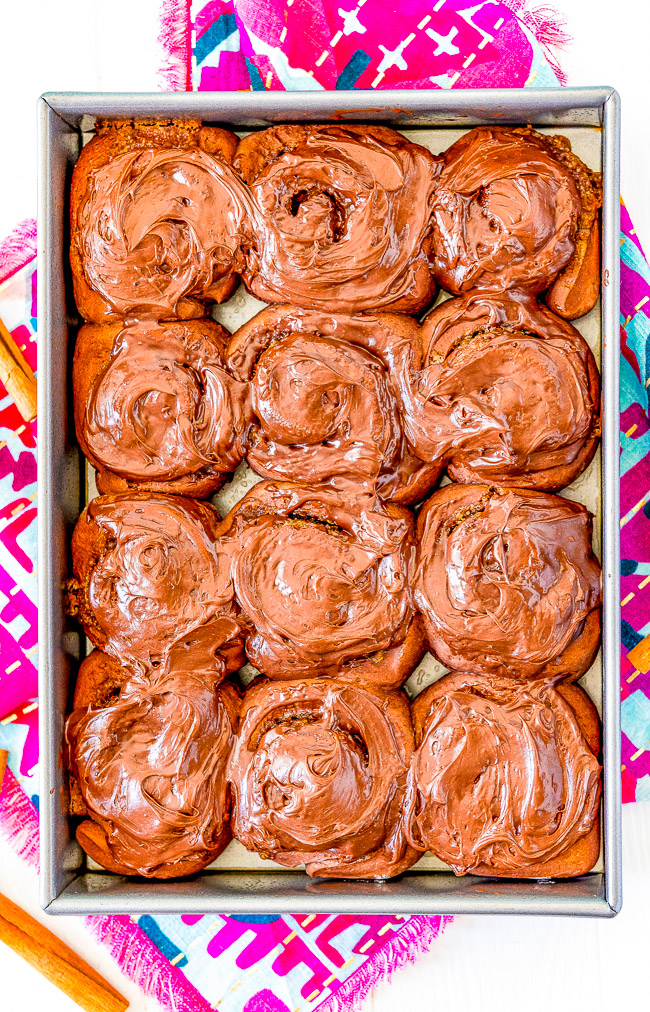 A baking tray with twelve chocolate-frosted cinnamon rolls on a colorful cloth background.