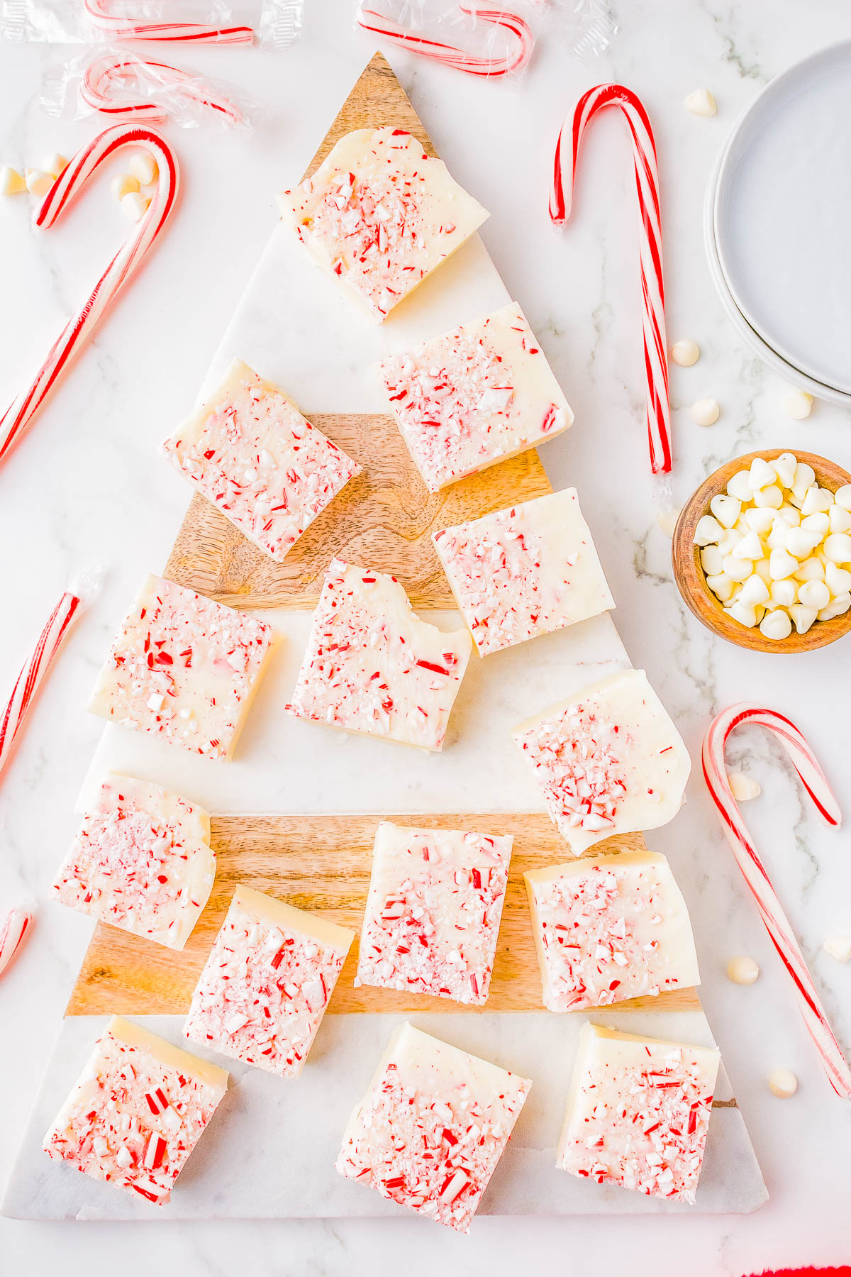 A wooden board shaped like a Christmas tree holds pieces of peppermint fudge. Candy canes, a bowl of marshmallows, and a plate surround the board on a marble surface.