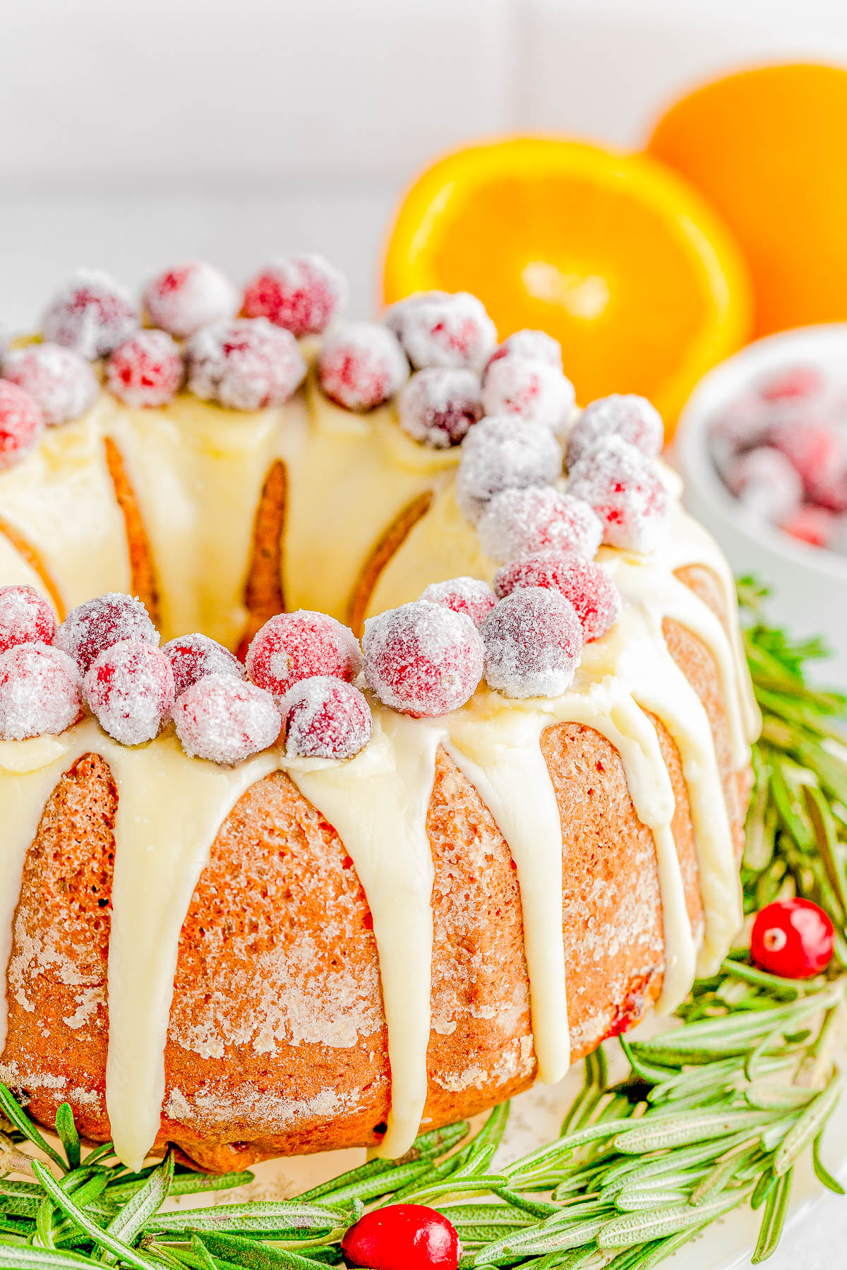 A Bundt cake coated with white icing and topped with sugared cranberries is surrounded by sprigs of rosemary. Orange halves and a bowl with more cranberries are in the background.