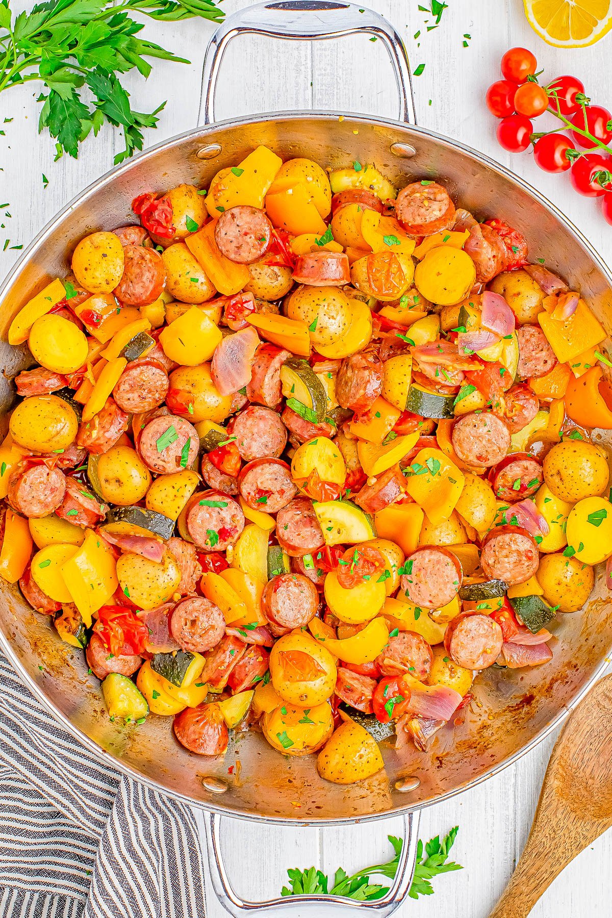 A skillet filled with sliced sausage, yellow bell peppers, potatoes, and tomatoes, garnished with parsley. A striped towel and fresh vegetables are nearby.