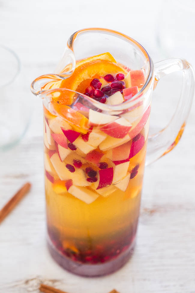 A clear pitcher filled with white sangria, containing sliced apples, oranges, and pomegranate seeds.