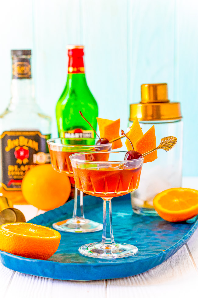 Two cocktails with orange and cherry garnishes on a blue tray, surrounded by a bottle of whiskey, a bottle of vermouth, a cocktail shaker, and fresh oranges.