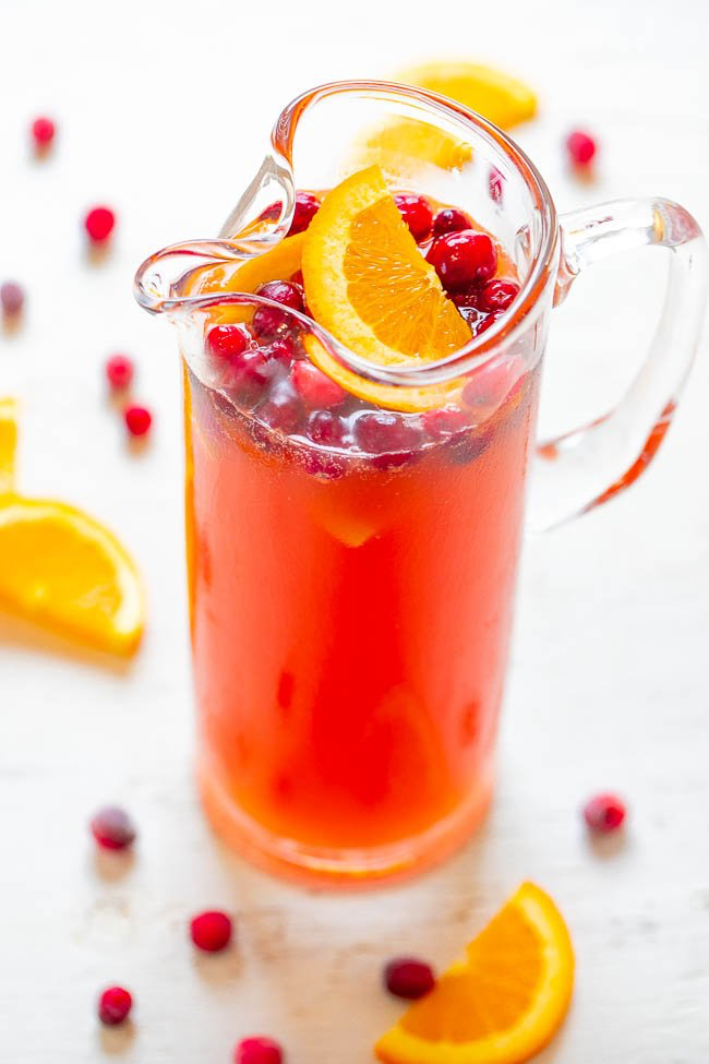 A glass pitcher filled with red punch, garnished with cranberries and orange slices. Orange wedges and cranberries are scattered around on the white surface.