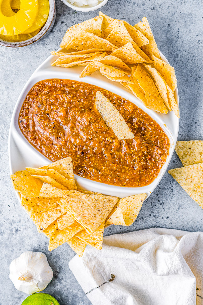 A white bowl filled with salsa and surrounded by tortilla chips on a gray surface, with a lime, garlic, pineapple, and cloth nearby.