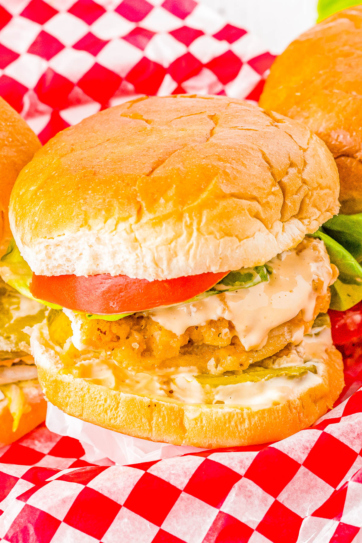 A chicken sandwich with lettuce, tomato, and sauce on a bun, placed on a red and white checkered paper.