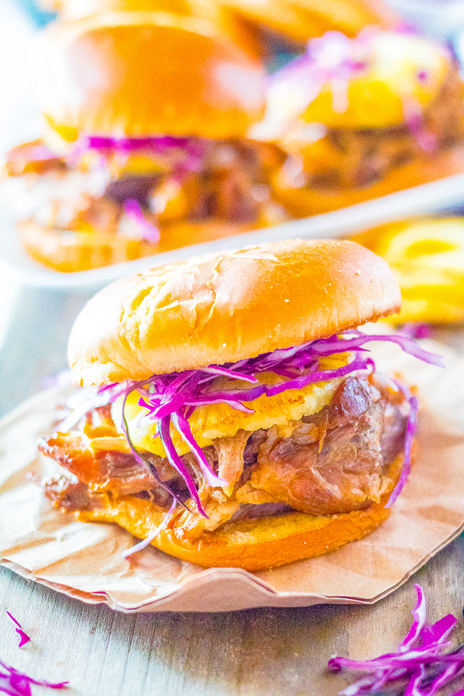 Pulled pork sandwich with red cabbage and pineapple on a bun, set on a brown napkin, with more sandwiches in the background.