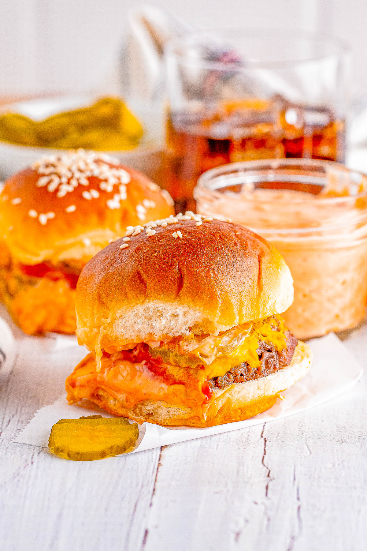 Two cheeseburgers with melted cheese on buns are placed next to a pickle slice. A jar of sauce and a glass of iced tea are in the background.