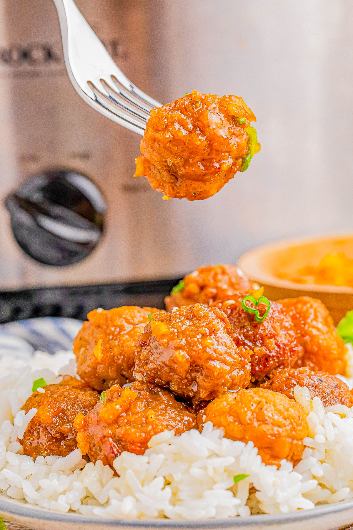 A fork holds a glazed meatball above a plate of rice topped with several glazed meatballs.