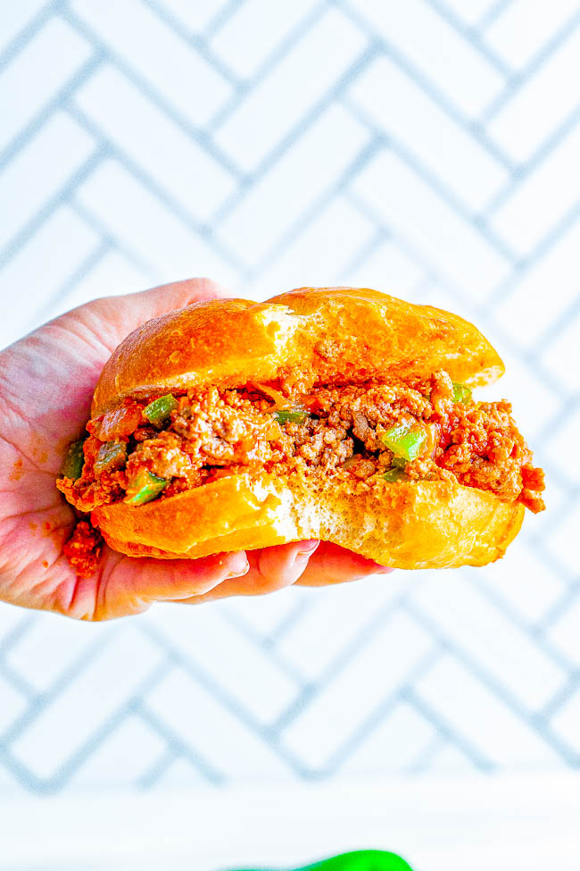 Close-up of a hand holding a sloppy joe sandwich with meat and peppers in a soft bun against a white, patterned background.