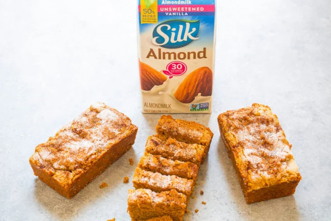 Three slices of cinnamon bread are arranged with a carton of Silk Almond Unsweetened Vanilla milk in the background on a light-colored surface.