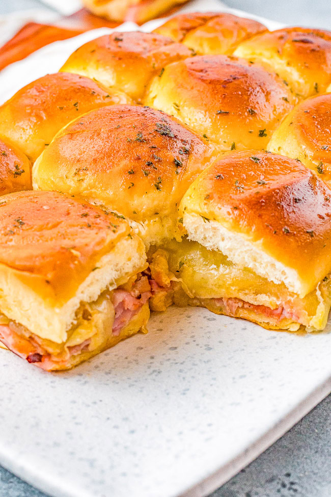 Close-up of baked ham and cheese sliders on a white plate, with one slider partially pulled out, displaying the filling.