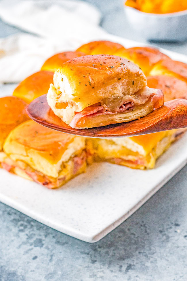 A wooden spatula holds a ham and cheese slider with a golden top above a plate of similar sliders on a gray surface.