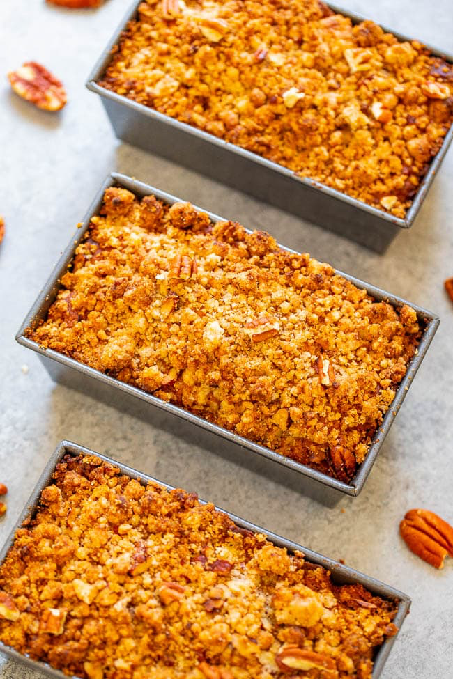 Three rectangular baking pans filled with pecan crumb-topped bread placed on a light-colored surface. Several pecan pieces are scattered around the pans.