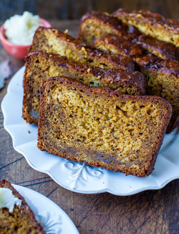 A white plate with several slices of banana bread neatly arranged, featuring a golden-brown crust and moist interior. In the background, there is a small bowl of butter.