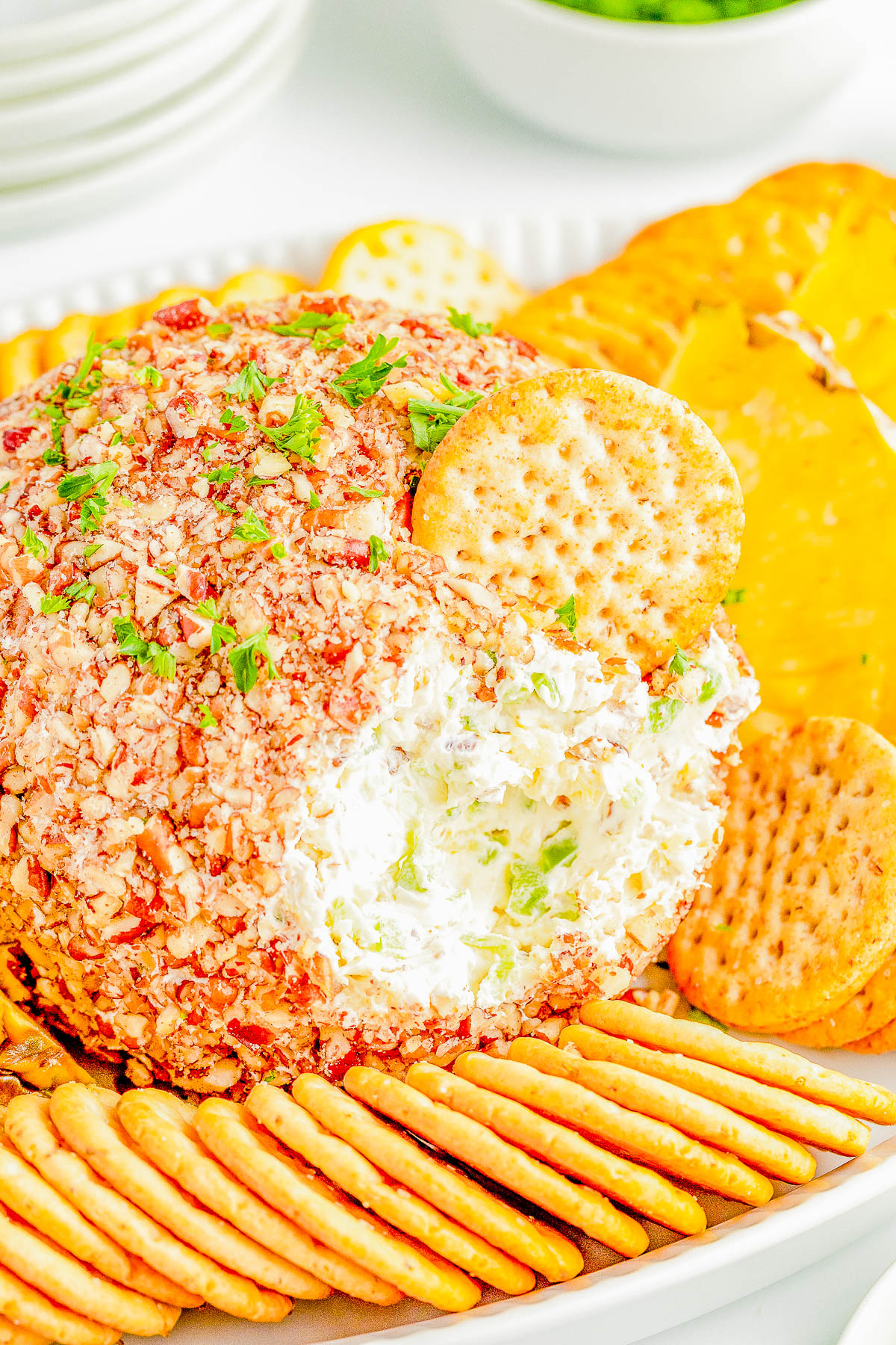 A cheese ball coated with chopped nuts, partially eaten, surrounded by assorted crackers and slices of yellow bell pepper on a white platter.