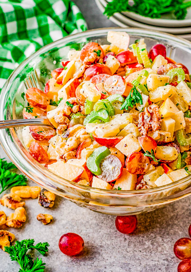 A glass bowl of Waldorf salad with apples, grapes, celery, and walnuts. A fork is placed in the bowl. Plates and greens are in the background.