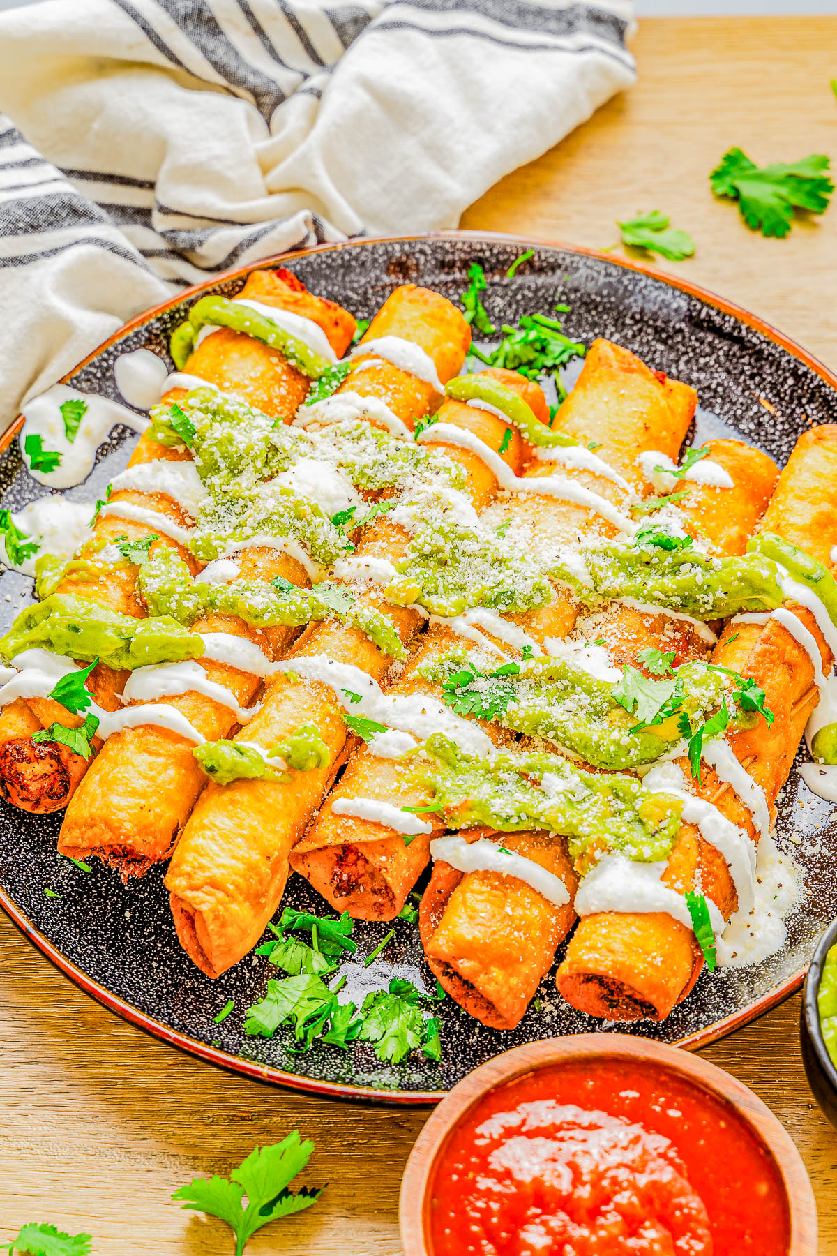 A plate of rolled, fried taquitos topped with green sauce, white drizzle, and cheese, garnished with cilantro. A small bowl of red sauce is nearby. A striped cloth is in the background.