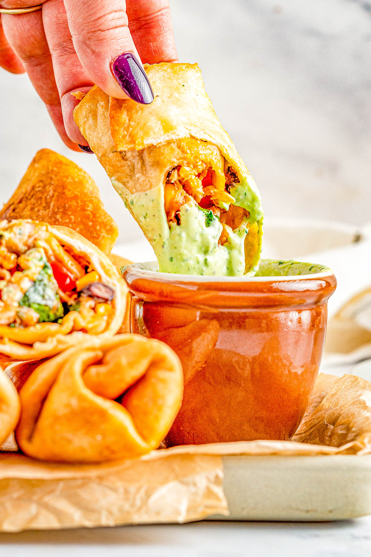 A hand dips a fried tortilla roll filled with vegetables into a green sauce in a brown cup. Other rolls are on a paper in the background.