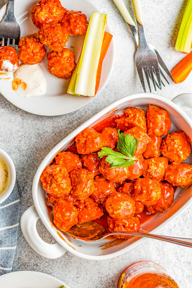 A white dish filled with sauce-covered meatballs garnished with parsley. Nearby, a small plate has celery, carrot sticks, and meatballs. Forks and dip bowls are placed around.